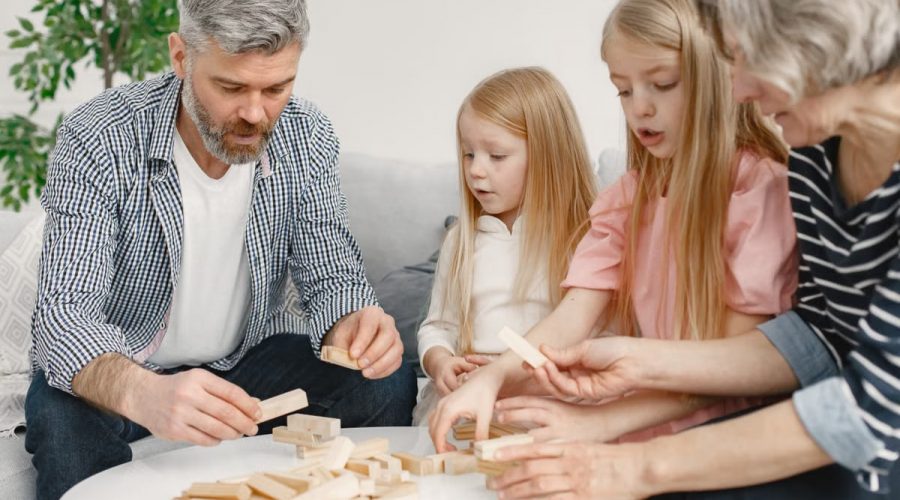 Four family members of different ages engage with toy blocks on a table indoors.